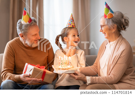 A young girl, wearing a festive party hat, gleefully receives a birthday cake from her grandmother, while an equally joyous grandfather looks on holding a gift A young girl, wearing a festive party hat, gleefully receives a birthday cake from her grandmother, while an equally joyous grandfather looks on holding a gift 114701814