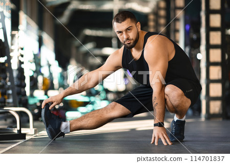 A man is squatting on the ground in a gym. He is wearing workout attire and appears focused on his workout routine. The gym is well-equipped with various exercise machines in the background. 114701837