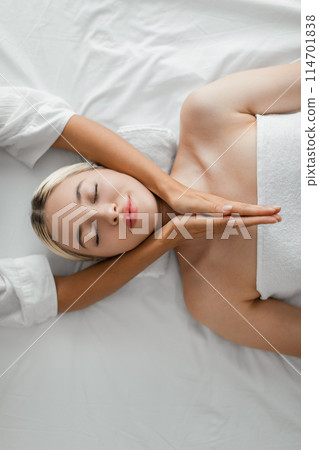Top view of young woman lies on her back with eyes closed, enjoying a relaxing facial massage from a pair of hands, indicating a sense of tranquility and self-care during a spa treatment. 114701838