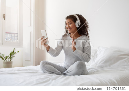 A cheerful young Hispanic woman sits cross-legged on a white bed, wearing cozy casual attire and headphones, engaged in a video call A cheerful young Hispanic woman sits cross-legged on a white bed, wearing cozy casual attire and headphones, engaged in a video call 114701861