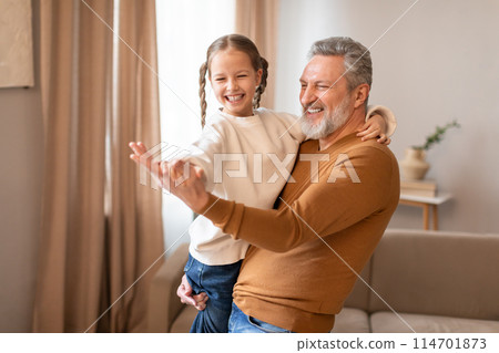 Senior man is standing, holding a little girl in his arms, grandfather dancing with his cute little granddaughter at home Senior man is standing, holding a little girl in his arms, grandfather dancing with his cute little granddaughter at home 114701873