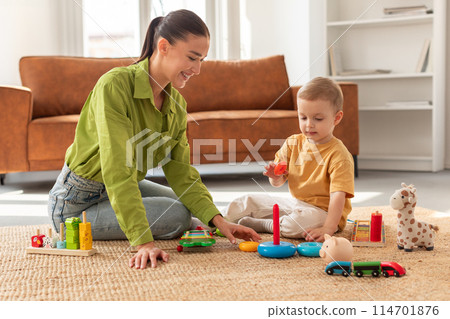 A woman is sitting on the floor, engaging in play with a child. The boy is smiling and appears to be enjoying the interaction. They are surrounded by toys and colorful objects. 114701876
