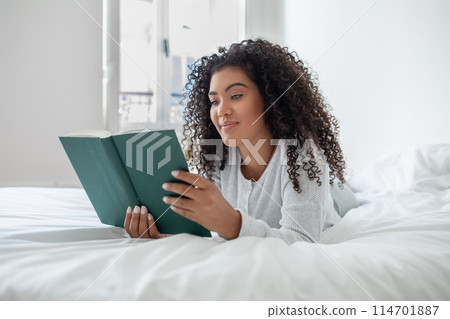 Hispanic woman is reclining on a bed with a book in her hands, absorbed in reading. The room is cozy and well-lit, creating a comfortable reading environment. 114701887