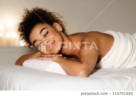 Close-up of a joyful african american woman with a radiant smile, wrapped in a towel in a spa environment 114701888