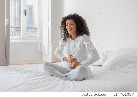 Young hispanic woman wearing pajamas sitting in bed with hands on abdomen, feeling unwell in the morning 114701897
