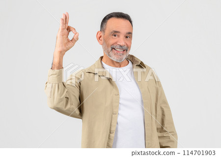 A senior man wearing a white shirt and tan jacket is seen making the Okay sign with his hand. His fingers are split in the iconic OK shape 114701903