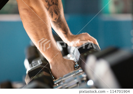 Cropped of man with a tattoo on his arm is lifting a pair of dumbbells with focused concentration, showcasing his strength and dedication to fitness Cropped of man with a tattoo on his arm is lifting a pair of dumbbells with focused concentration, showcasing his strength and dedication to fitness 114701904