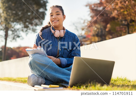 A girl student is sitting on the ground outdoors, working on a laptop, doing homework, smiling at camera, copy space 114701911