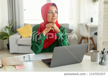 Muslim woman is seated at a table, concentrating on her laptop screen. She appears focused and engaged in her work or study, surrounded by a simple and modern workspace, copy space Muslim woman is seated at a table, concentrating on her laptop screen. She appears focused and engaged in her work or study, surrounded by a simple and modern workspace, copy space 114701937