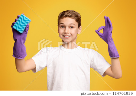 Housekeeping concept. Cute boy in cleaning gloves showing OK sign and holding sponge, orange panorama background 114701939