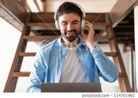 A man is sitting with headphones on, focused on a laptop. He is typing on the keyboard, with a serious expression on his face. The man seems engrossed in his work or studies 114701962