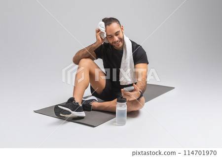 A man is seated on a floor mat with a towel draped around his neck. He appears relaxed and may have just finished exercising or working out, checking smartphone A man is seated on a floor mat with a towel draped around his neck. He appears relaxed and may have just finished exercising or working out, checking smartphone 114701970