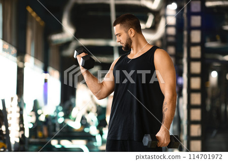 A toned man with a well-defined physique is engaged in a workout session, performing bicep curls with a dumbbell at modern gym 114701972