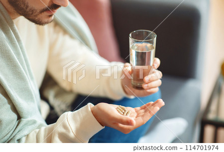 Guy is seated comfortably on a couch, holding a glass of water in one hand and a handful of pills in the other, likely preparing to take medication 114701974