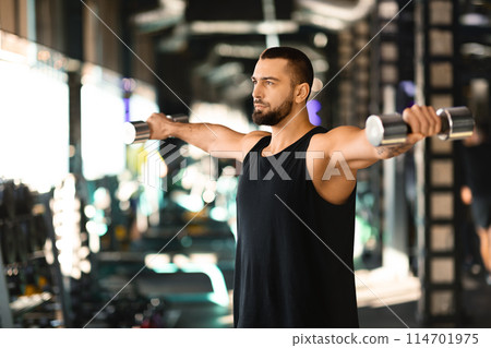 A man is shown in a gym holding a barbell, showcasing his strength and dedication to fitness. He is focused and determined as he lifts the heavy weight, surrounded by exercise equipment. 114701975