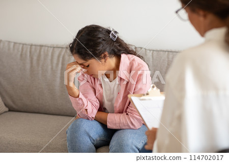 A young woman sits on a couch, her body language expressing distress as she covers her face with her hand. In the foreground, a therapist, indicated by the presence of a clipboard, offers a support 114702017