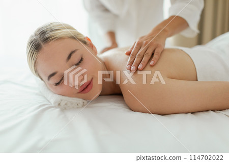 A woman lying face down on a massage table at a luxury spa while a massage therapist is massaging her upper back. The room is dimly lit with soothing music playing in the background. 114702022