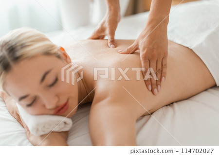 A woman is lying face down on a massage table, receiving a back massage from a massage therapist in a cozy spa setting. The therapists hands are shown applying pressure to the woman back muscles. 114702027