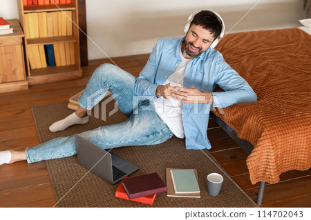 A man is sitting on the floor with his laptop open in front of him, appearing focused on smartphone, working from home, high angle view 114702043