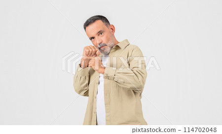 A senior man wearing a tan shirt is standing while holding his hands together in front of him in fighting pose. He appears focused and contemplative as he holds his hands clasped together. A senior man wearing a tan shirt is standing while holding his hands together in front of him in fighting pose. He appears focused and contemplative as he holds his hands clasped together. 114702048