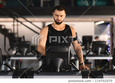 A man wearing athletic clothing is jogging on a treadmill machine inside a bright and modern gym. The equipment is surrounded by other exercise machines and gym-goers in the background. 114702070