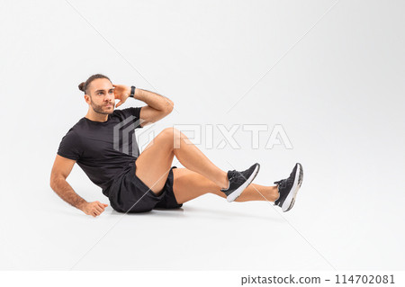 A man wearing a black shirt and shorts is seated on the ground, doing workout crunches, exercising on white studio background A man wearing a black shirt and shorts is seated on the ground, doing workout crunches, exercising on white studio background 114702081