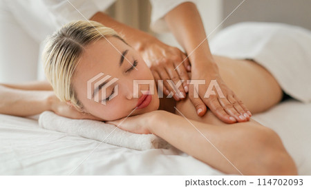 A woman is lying face down on a massage table while a massage therapist is applying pressure to her shoulders muscles. The woman appears relaxed and comfortable as she enjoys the soothing massage. A woman is lying face down on a massage table while a massage therapist is applying pressure to her shoulders muscles. The woman appears relaxed and comfortable as she enjoys the soothing massage. 114702093