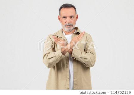 A senior man is shown standing upright with his hands folded in front of his chest in a confident and attentive posture, showing stop gesture on white 114702139