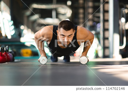A man is performing push ups while holding two dumbbells in his hands. He is engaging his chest, triceps, and core muscles in this strength training exercise. 114702146
