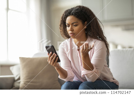 This image shows an African American woman on her couch, frowning at her phone, surrounded by a comfortable home environment, depicting a moment of stress 114702147