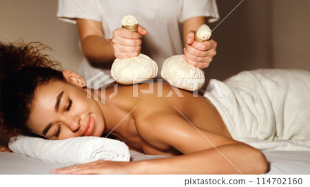 African american woman lies face down, receiving a traditional herbal compress massage from a masseuse in a serene spa setting African american woman lies face down, receiving a traditional herbal compress massage from a masseuse in a serene spa setting 114702160