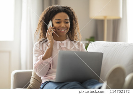 Captured in a cozy living room, this photo shows a focused African American woman talking on the phone while working on her laptop, embodying a productive home office setting Captured in a cozy living room, this photo shows a focused African American woman talking on the phone while working on her laptop, embodying a productive home office setting 114702172