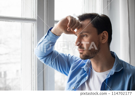 A man is standing by a window, looking outside. His hand is resting on his head as he appears lost in thought, perhaps contemplating or reflecting A man is standing by a window, looking outside. His hand is resting on his head as he appears lost in thought, perhaps contemplating or reflecting 114702177