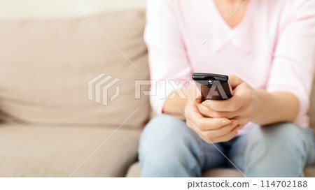 Lady is seated on a comfortable beige couch with hands wrapped around a television remote control, seemingly ready to change channels or adjust the volume while enjoying some leisure time at home. 114702188