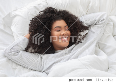 Hispanic woman is lying in bed with her head resting on her hands, looking contemplative and relaxed in the morning. The room is dimly lit, emphasizing her peaceful expression. Hispanic woman is lying in bed with her head resting on her hands, looking contemplative and relaxed in the morning. The room is dimly lit, emphasizing her peaceful expression. 114702198