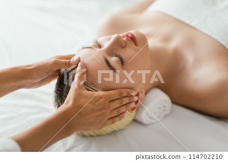 A woman is lying down with her eyes closed, receiving a relaxing facial massage. The therapists hands are gently massaging her face, view above 114702210