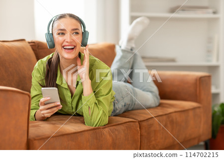 A woman is lying comfortably on a couch with headphones covering her ears, holding smartphone. She appears relaxed and engaged in listening to music or an audio program. 114702215