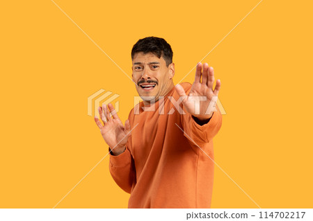 Frightened man is seen standing in front of a bright yellow background, outstretching hands. He appears relaxed and confident, wearing casual clothing. The yellow backdrop 114702217