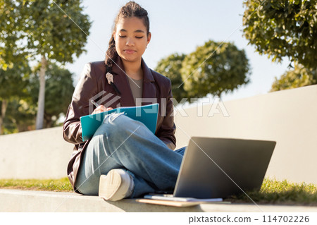 A focused young girl is sitting on a concrete step outdoors, writing on a clipboard with her laptop open beside her. She is casually dressed, enjoying the warm sunshine, studying in a park setting. 114702226
