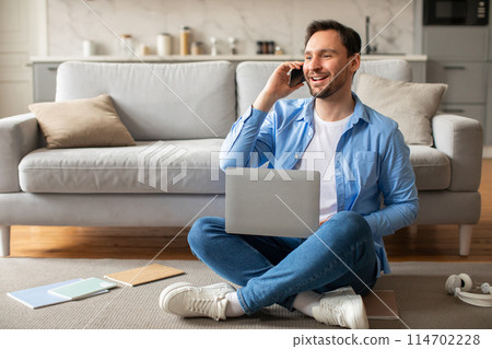 A man is seated on the floor, multitasking by using a laptop and talking on a cell phone. He appears focused and engaged in his work, balancing technology use efficiently. A man is seated on the floor, multitasking by using a laptop and talking on a cell phone. He appears focused and engaged in his work, balancing technology use efficiently. 114702228