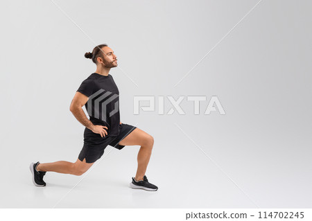 A man with a beard and bun hair style is captured mid-motion while performing a lunge. He is wearing a black t-shirt, black shorts, and athletic shoes, copy space A man with a beard and bun hair style is captured mid-motion while performing a lunge. He is wearing a black t-shirt, black shorts, and athletic shoes, copy space 114702245