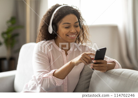 This image portrays a content African American woman relaxing on her sofa, enjoying music through her headphones and phone, creating a tranquil atmosphere in a modern home environment This image portrays a content African American woman relaxing on her sofa, enjoying music through her headphones and phone, creating a tranquil atmosphere in a modern home environment 114702246