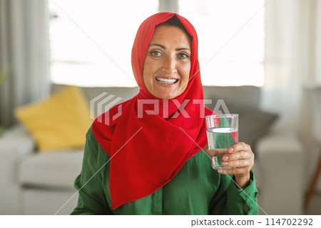 A woman wearing a red and green hijab is holding a glass of water in her hands. She is standing indoors, and the focus is on her colorful head covering and the clear glass of water she is holding. A woman wearing a red and green hijab is holding a glass of water in her hands. She is standing indoors, and the focus is on her colorful head covering and the clear glass of water she is holding. 114702292