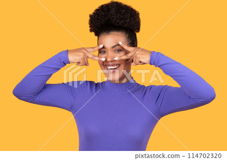 Hispanic woman standing and forming peace gesture with her hands in front of her eyes. She is wearing a casual outfit and appears to be smiling, yellow background Hispanic woman standing and forming peace gesture with her hands in front of her eyes. She is wearing a casual outfit and appears to be smiling, yellow background 114702320