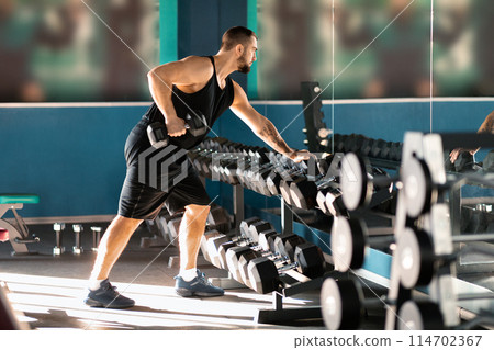 A man in athletic attire is seen working out with a rack of dumbbells at a gym. He is lifting weights with focused determination, showcasing strength training as a key part of his fitness routine. 114702367