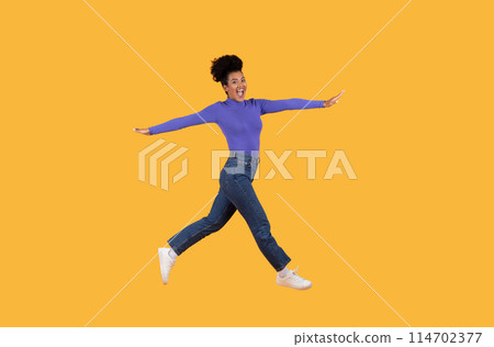 A woman with a purple shirt is energetically jumping in the air, showcasing a burst of movement and joy, isolated on yellow studio background 114702377