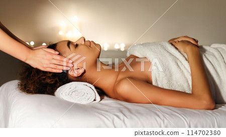 An African American woman enjoys a calming spa treatment performed by a professional therapist, promoting wellness and relaxation 114702380