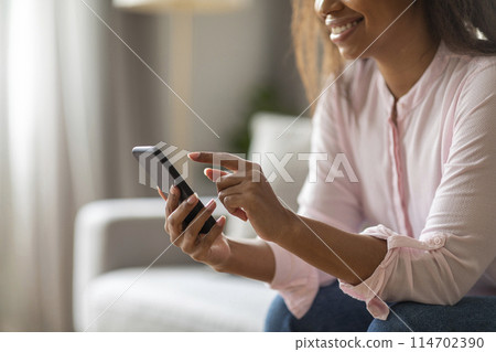 Enjoying a quiet day indoors, black woman sits on her couch using her mobile phone, surrounded by a tastefully decorated living room in soft colors, cropped Enjoying a quiet day indoors, black woman sits on her couch using her mobile phone, surrounded by a tastefully decorated living room in soft colors, cropped 114702390