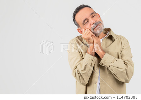 A senior man is standing with his hands pressed together against his face, feeling sleepy on white background, copy space 114702393
