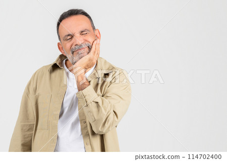 A senior man sits with his hand placed on his face, suffering from toothache on white background, copy space 114702400